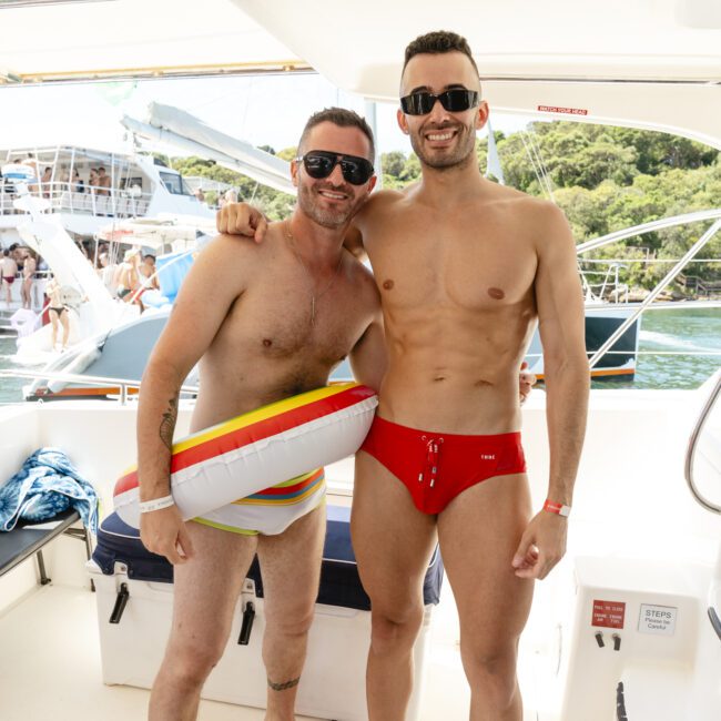 Two men pose on a boat, both in swimwear. One wears shorts and a colorful float ring, and the other in red swim trunks. They're smiling, with a sunny background featuring other boats and lush greenery.