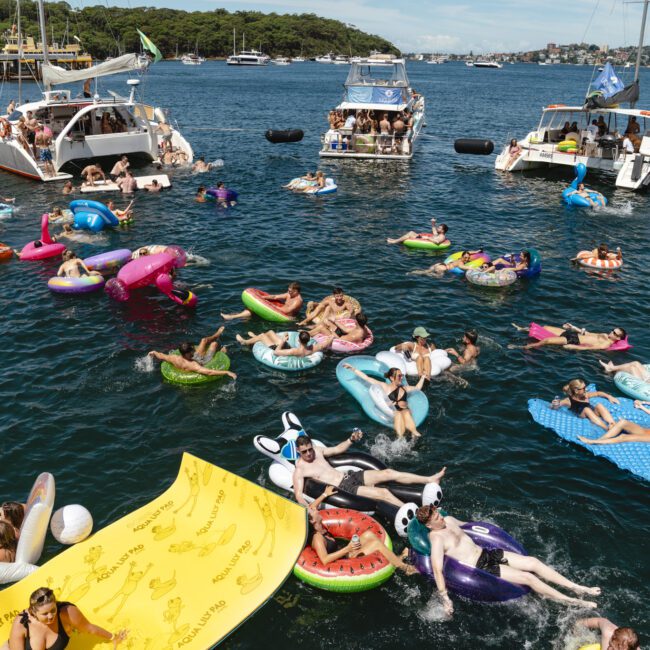 People are enjoying a sunny day on colorful inflatable floats in a bay. Several boats are anchored nearby, with more people on board. The scene is lively, with clear blue water and a forested shoreline in the background.