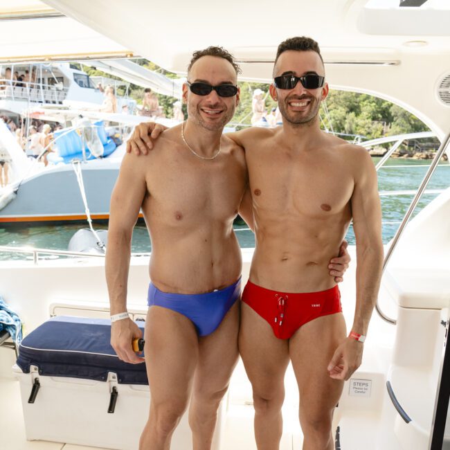 Two men wearing sunglasses and swim briefs, one in blue and the other in red, stand arm in arm on a boat. They are smiling at the camera. A scenic view with trees and water is visible in the background.