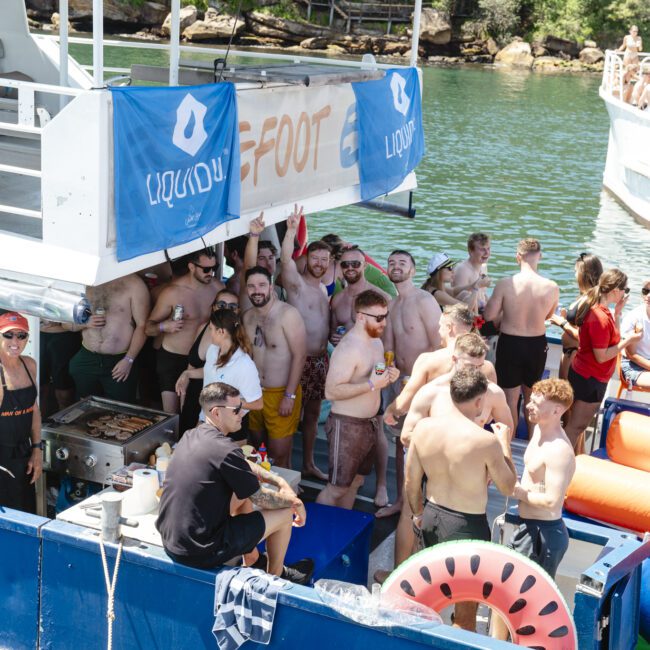 A group of people are gathered on a blue raft with inflatable floats, enjoying a sunny day on the water. Many are wearing swimsuits and sunglasses. A boat is docked nearby, and lush greenery is visible in the background.