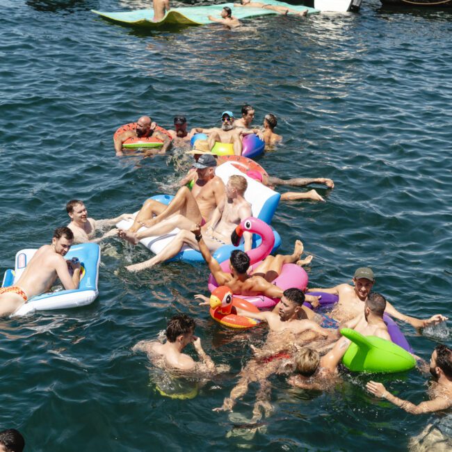 People enjoy a sunny day on a lake, relaxing on colorful inflatables and swimming near a boat. The scene is lively with various floats shaped like a flamingo and other designs, surrounded by clear, calm water.