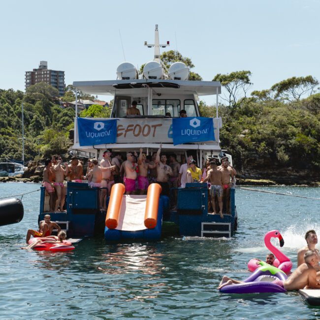 A group of people enjoys a sunny day on a boat with inflatable slides leading into the water. Some people are lounging on inflatable floats, including a flamingo, in a scenic water setting with trees and a building in the background.