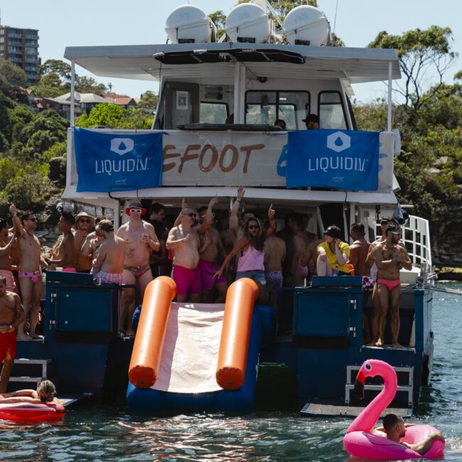 A group of people in swimsuits are partying on a boat named "Big Foot," featuring two water slides. Inflatable pool toys, including a flamingo, float around them in the water under a bright, sunny sky. Forested shorelines are in the background.