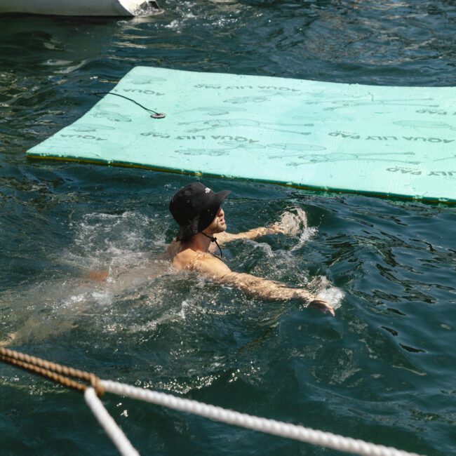 A person swimming in a body of water near a docked boat and a large floating green mat. The swimmer is wearing a black swim cap and goggles and is making a splash around them. Ropes from the dock are partially visible in the foreground.