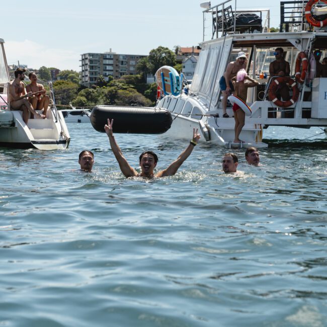 A group of people swimming in a sunny harbor near two boats. One person in the foreground raises arms in a celebratory gesture. The boat on the right has "Rum Runner Cruises" written on it. Trees and buildings are visible in the background.