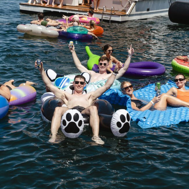 A group of people enjoying a sunny day on the water, lounging on inflatable floats near a boat. Two men on a large float raise their drinks and hands in celebration. Others relax on various colorful floats nearby.