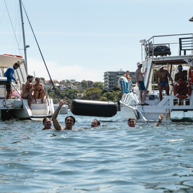 A group of people swim in the sea between two anchored boats on a sunny day. They are smiling and holding drinks, with others watching from the boats. Buildings and greenery are visible in the background.