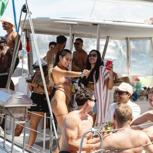 A group of people enjoying a sunny day on a boat, wearing swimsuits and summer attire. Some hold drinks and engage in lively conversation. The sky is clear, and a flag is visible in the background. The atmosphere is festive and relaxed.
