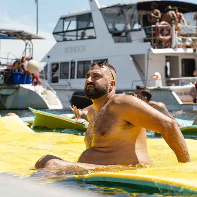 A man sits on a large yellow float in the water, surrounded by several people enjoying the sunny day. Behind him, two boats are anchored, with more people on the decks. The sky is clear and bright.