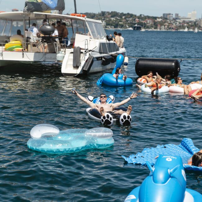People are relaxing on colorful inflatable floats in a sunny waterway, with a sailboat nearby. The scene includes various floats like a swan and a panda, as well as people sunbathing and enjoying the water. A cityscape is visible in the background.