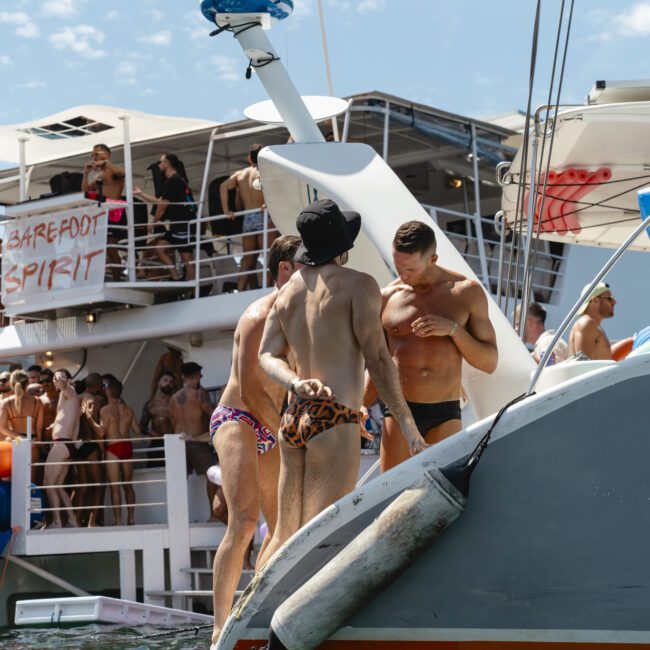 A crowd on a boat enjoying a sunny day. People are socializing and relaxing, with some standing on an adjacent boat. A “Barefoot Spirit” sign is visible on the main boat. The atmosphere suggests a lively party or gathering on the water.