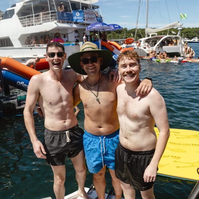Three men in swim trunks smile and pose on a boat dock. Behind them, boats and people enjoying a sunny day on the water are visible. One man wears sunglasses and a wide-brimmed hat. Inflatable water toys float nearby.