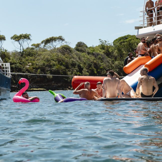 People in swimsuits enjoying a party on a lake with inflatable toys, including a pink flamingo and a unicorn. Multiple boats are tied together, and some guests are using a water slide from one of the boats. Trees are visible in the background.