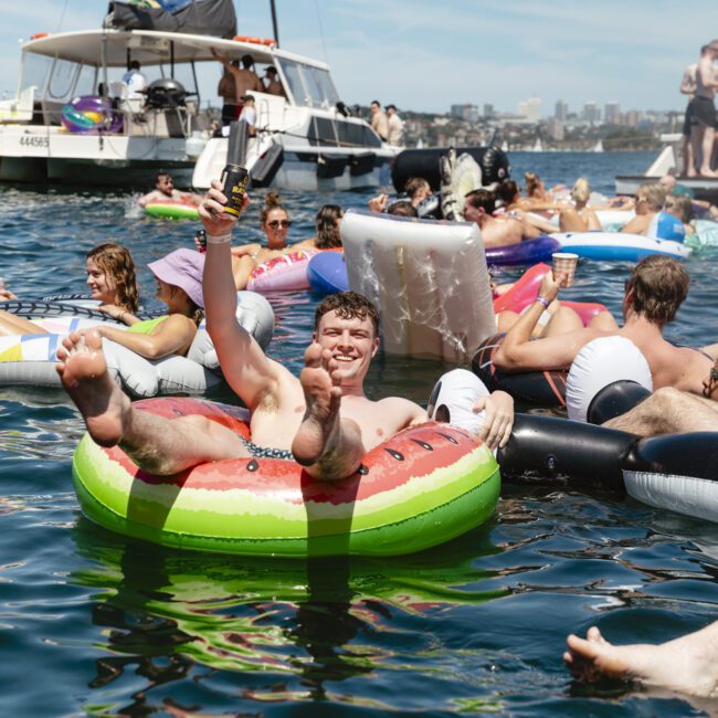 A group of people enjoying a sunny day on the water with inflatables of various shapes and colors. Some are holding drinks, and boats are visible in the background. Everyone appears relaxed and happy.