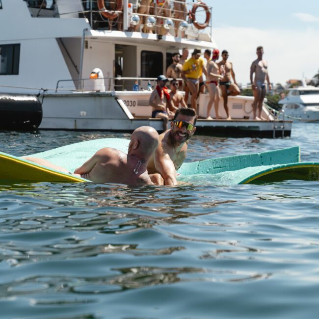 People enjoying the water near a boat. Two individuals play on floating mats while others gather on the boat in the background. The scene is lively, with a clear sky and other boats visible.