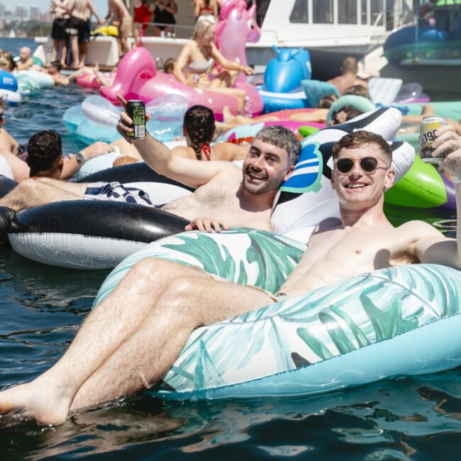 Two smiling men float on inflatable pool toys in a crowded body of water. One holds a drink and wears sunglasses. Other people on colorful inflatables are seen in the background, with a large boat nearby under a sunny sky.