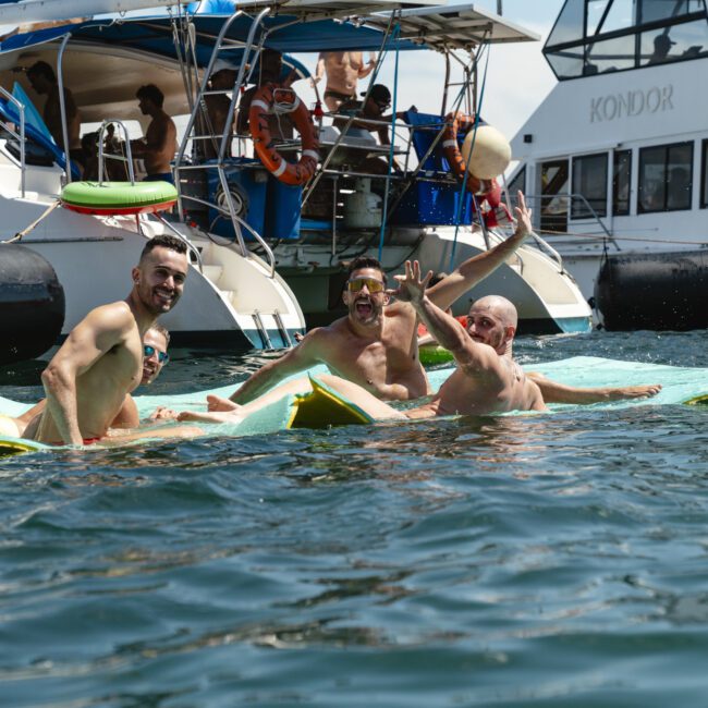 Four men laughing and playing with a ball while lounging on floating mats in the water. Two boats are anchored nearby, and the weather is sunny with clear skies.