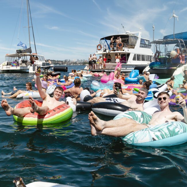 A group of people enjoying a sunny day on a lake, lounging on colorful inflatable floats near several boats. They are socializing and waving at the camera, with a clear blue sky above them.