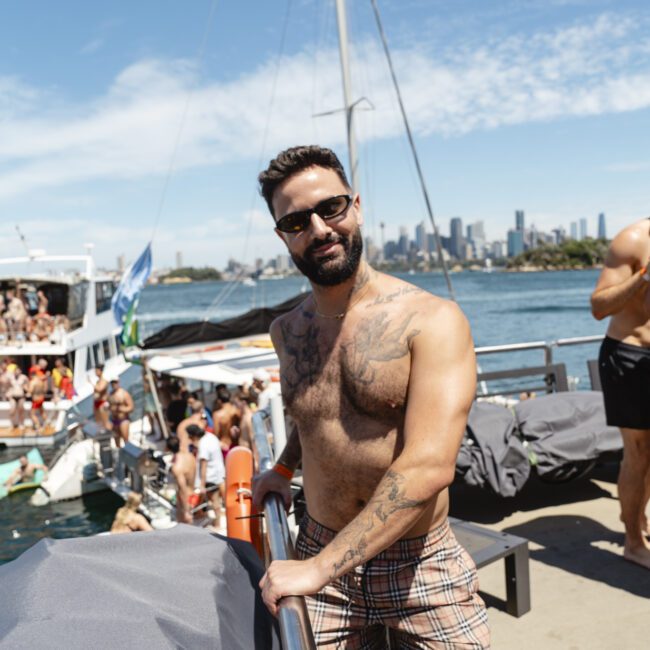 A man wearing sunglasses and plaid shorts stands on a boat, looking at the camera. He has tattoos on his chest and arm. Behind him, people are socializing on a boat, with a city skyline in the background under a clear blue sky.