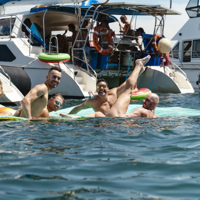 Four people in swimwear are joyfully lounging on a float in the water, with yachts anchored in the background. They appear to be having fun, with one person playfully lifting a leg in the air. The scene is bright and sunny.