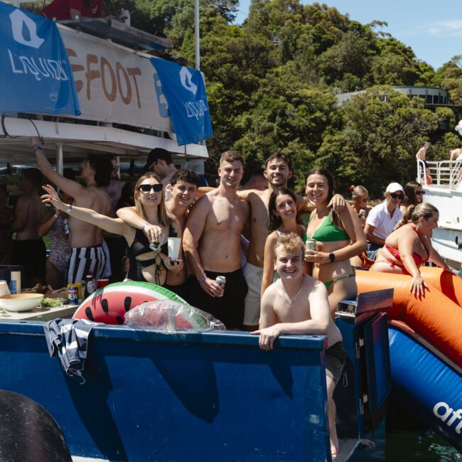 A group of people in swimwear smiles and poses on a boat with a banner reading "Lazy Foot." Surroundings include trees and water. One person holds an inflatable watermelon ring. The atmosphere appears relaxed and festive.