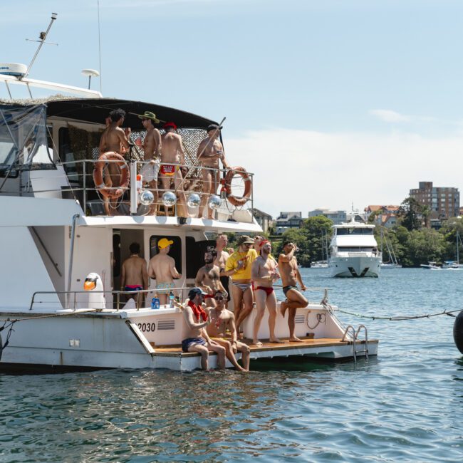 A group of people enjoying a sunny day on a yacht. They are gathered on the deck and some sit on the edge with feet near the water. In the background, buildings and trees line the shore. Another boat is visible on the water.