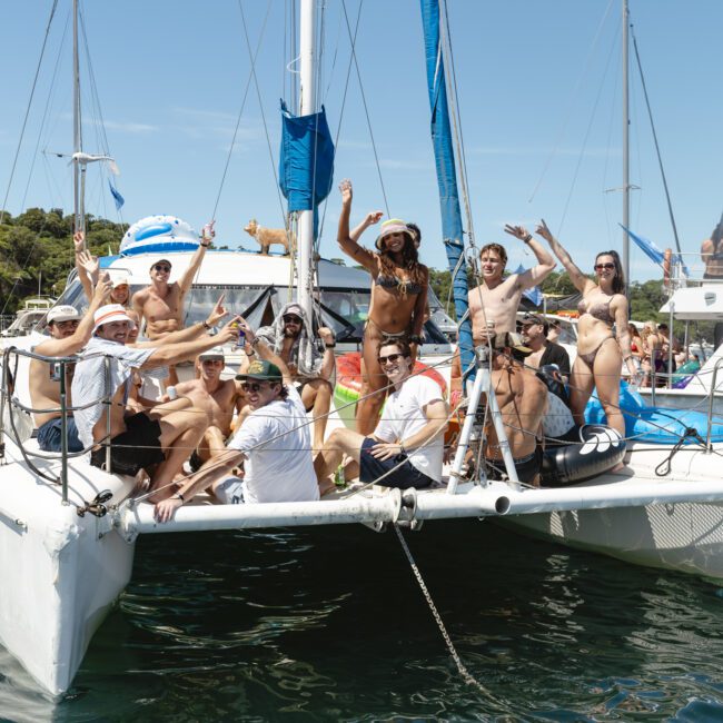 A group of people enjoying themselves on a catamaran in a sunny marina. They are smiling, waving, and wearing swimwear, with several boats visible in the background. The water is calm, and trees are lining the shore.