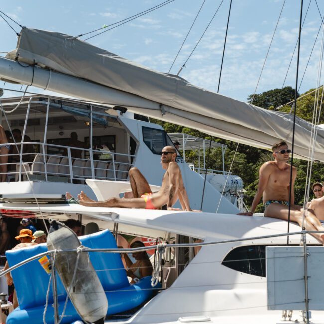 People relaxing on a sailboat under a clear blue sky. Some are sunbathing, while others are standing or sitting and watching the scenery. Trees are visible in the background, and various items and equipment can be seen on the boat.