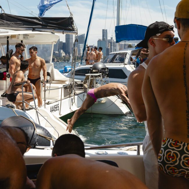 A group of people is enjoying a sunny day on a boat in the water. Some are wearing swimwear, and there are men diving and socializing. The skyline of a city is visible in the background.