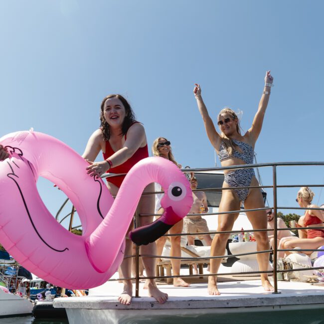 People on a boat deck under a clear sky. One person holds a pink inflatable flamingo, while another stands with arms raised. Others are sitting casually. Several boats are visible in the background.