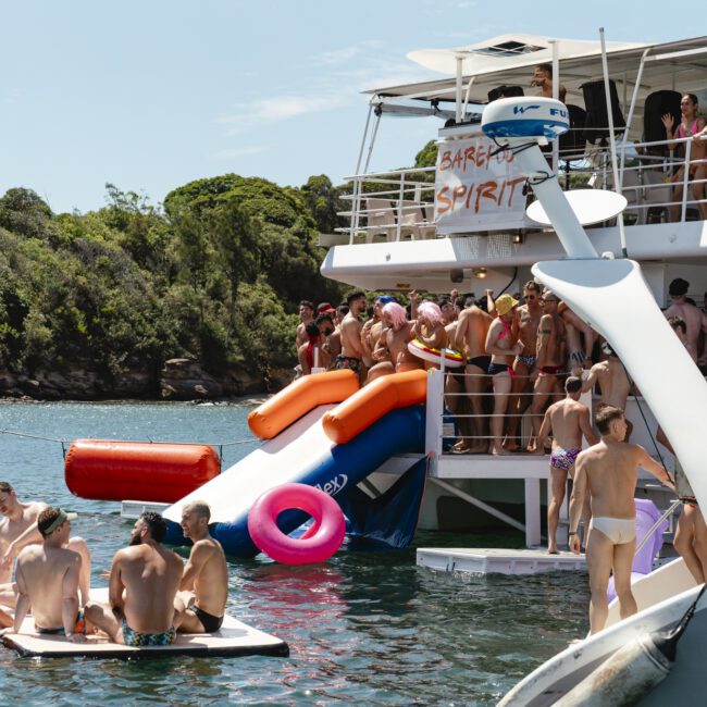 A lively boat party with people in swimwear enjoying the sun. Some are on the boat, while others are in the water with inflatables. A red and blue slide leads from the boat to the water. Trees and shoreline are visible in the background.