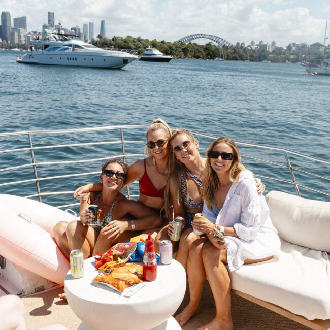 Four people relaxing on a boat, smiling and holding drinks. The scene includes a table with snacks and drinks. In the background, there is a city skyline, a large yacht, and a bridge over blue water. The sky is partly cloudy and sunny.