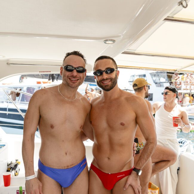 Two men in swim trunks, one in blue and the other in red, smiling on a boat. They are under a canopy with people and other boats visible in the background. The setting suggests a sunny day on the water.