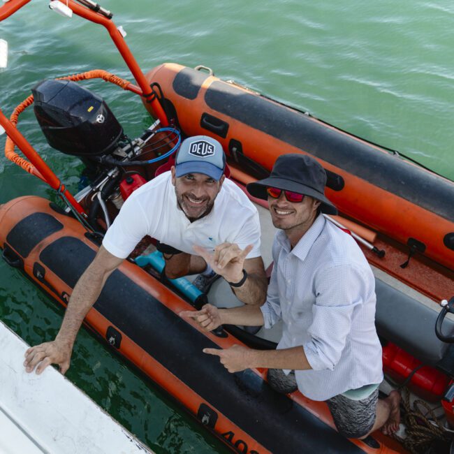 Two people on an orange and black inflatable boat on the water. One wears a cap and holds a drone controller, smiling, while the other, in a wide-brim hat and red sunglasses, points at a drone in hand. They are next to a white vessel.