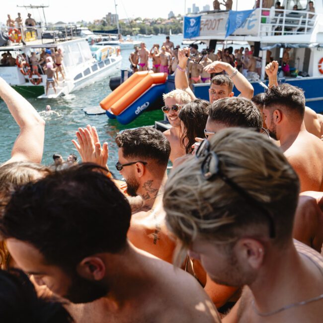 A large group of people are gathered closely, dancing and celebrating on a boat. Many are in swimwear, enjoying sunny weather. Other boats are visible in the background, with more people participating in the festivities on the water.