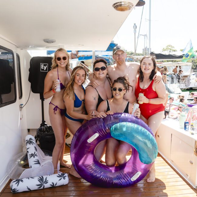 A group of six people smiling and posing on a boat deck. They are wearing swimwear and holding drinks and inflatable pool toys. Background features other boats and a clear, sunny sky.