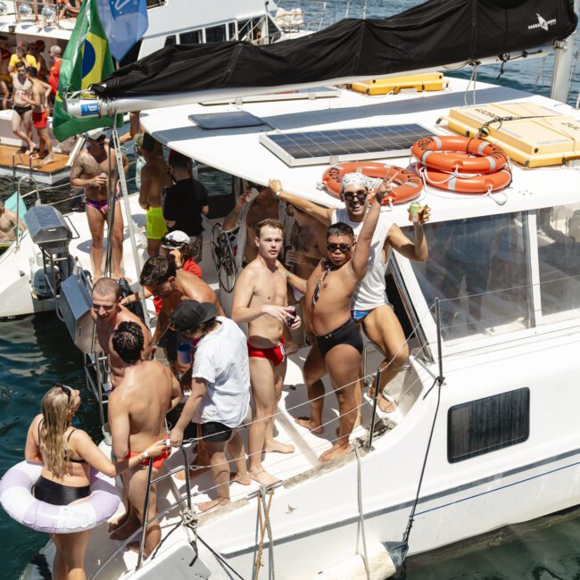 A group of people in swimwear enjoy a sunny day on a boat docked on a body of water. Some people are holding drinks and laughing. Flags and life preservers are visible. Other boats and city buildings are in the background.