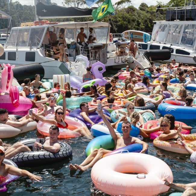 A large group of people relaxing on colorful inflatable tubes in the water, surrounded by boats. The scene is lively, with plenty of sunshine and people enjoying the day. A flag with a green, yellow, and blue design is visible in the background.