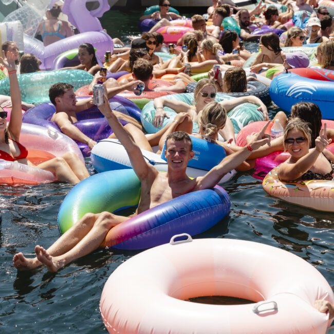 A group of people enjoying a sunny day on the water, lounging on colorful inflatable rings. They are smiling, raising drinks, and surrounded by boats. The scene is lively and festive, suggesting a fun gathering or party.