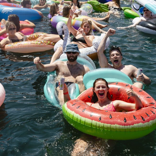 A group of people on colorful inflatable rafts relax and enjoy a sunny day on the water. The scene is lively, with smiles and drinks in hand, as many others float in the background.