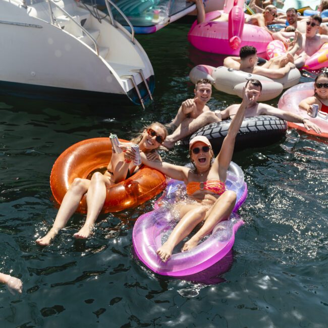 People enjoying a lively boat party, relaxing on colorful inflatable floaties in the water. They smile and raise drinks, surrounded by others on floats near a boat. The scene is festive and sunny.