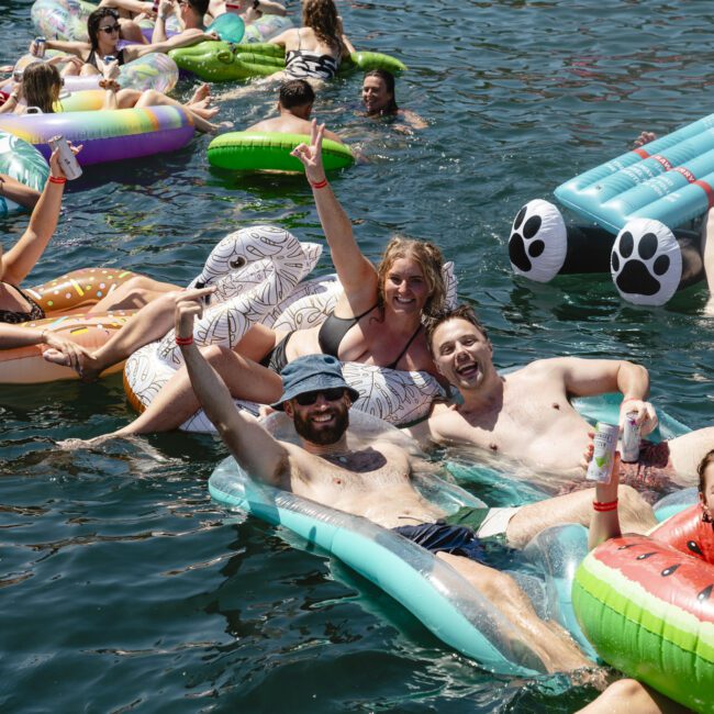 A group of people relaxing on colorful inflatables in a lake. They are smiling, raising their arms, and holding drinks. Bright sunshine illuminates the scene, highlighting the festive mood and variety of the pool floats.