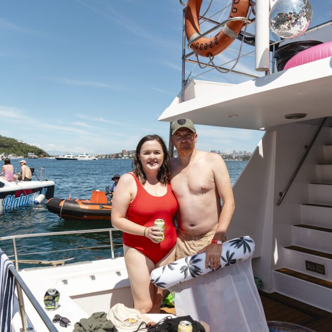 A person in a red swimsuit stands next to someone in a hat and shorts on a boat deck. They are holding drinks. In the background, there is water, a boat named "Palm Bay," and a glimpse of land. A lifebuoy and disco ball are visible on the boat.