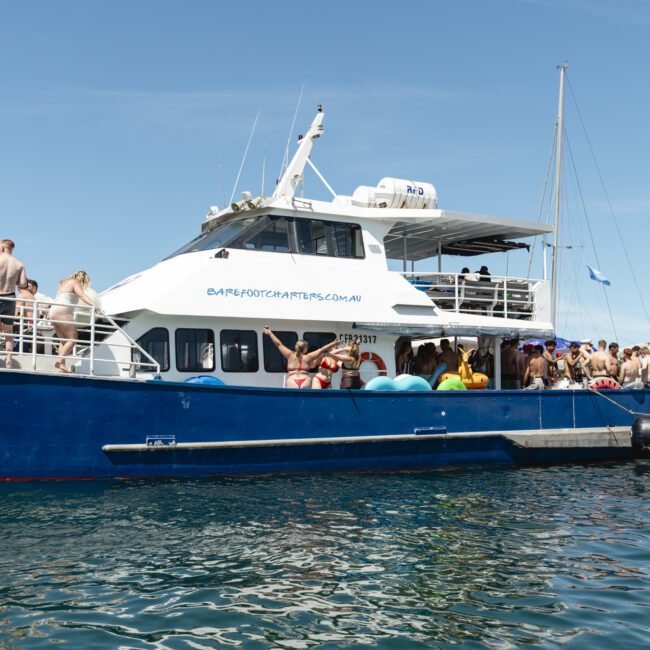 A large blue and white party boat with groups of people on board, some in swimwear, is anchored on a sunny day. The water is calm, and the sky is clear. Floaties are visible, and people are on the lower and upper decks.