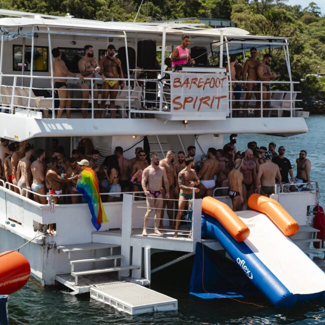 A boat packed with people in swimwear is on the water. Two inflatable slides descend into the water from the boat. A rainbow flag is draped over the side. Trees are visible in the background. A sign reads “Barefoot Spirit.”.