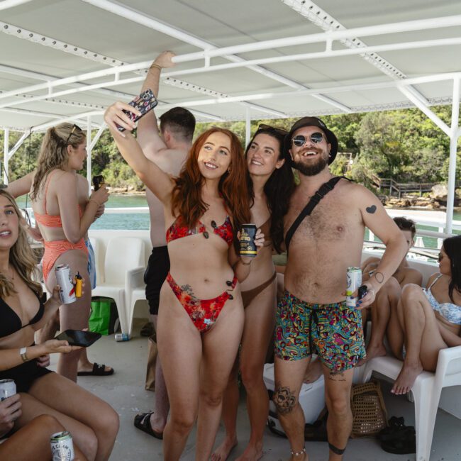 Group of people on a boat, wearing swimsuits. Three people in the center are smiling and holding drinks. Sunny day with greenery and water in the background.