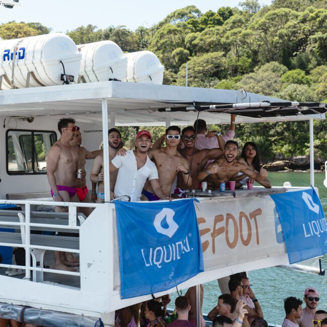 A group of people on a boat, many shirtless, smiling and waving at the camera. The boat has a banner with "LIQUI..." visible. Trees and water are in the background, suggesting a sunny day on the water.