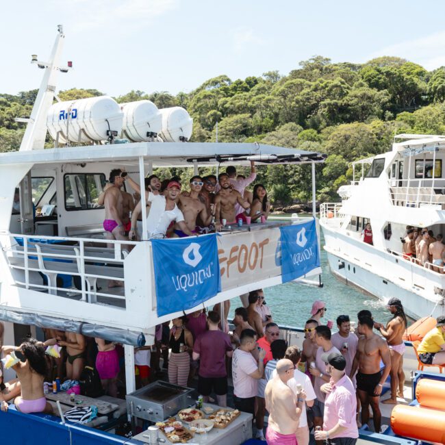 Group of people in swimwear enjoying a party on the deck of a boat. The boat is decorated with a banner reading "FOOT," and there's another banner with the word "liquid." Other people are mingling and socializing near tables of food.