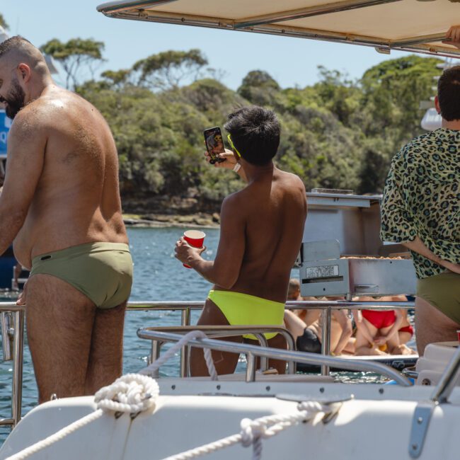 People on a boat are enjoying a sunny day. One person in green swimwear is taking a selfie, while others are relaxing. The background shows water and trees. The boat has the word "Voyager" visible.