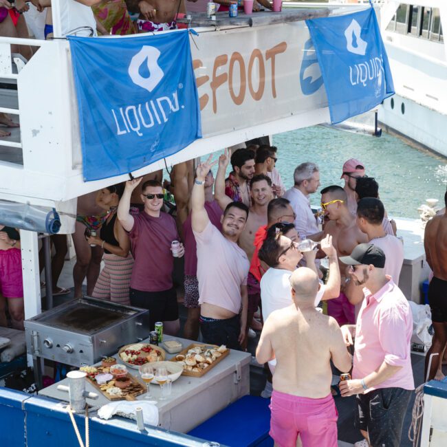 A lively group of people in pink and colorful outfits enjoy a party on a boat. The upper deck has a "Liquify" banner. Food is displayed on a table, and the boat is docked by the water. Some are dancing and raising their hands.